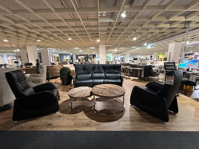 Trapezsofa mit zwei schwarzen Cumuly-Sesseln und runder Ablage in einem Möbelhaus.