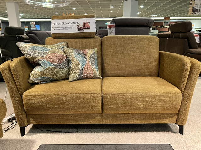Sofa Garnitur Alice in Senfgelb mit bunten Kissen, ohne Dekoelemente, ausgestellt im Möbelhaus.