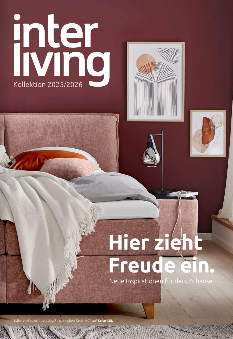 Interliving Wohnbuch 2026 Wohnzimmer mit einem beigen Sofa, dekorativen Kissen und einem Nachttisch mit Lampe. An der Wand hängen zwei Bilder. Text...